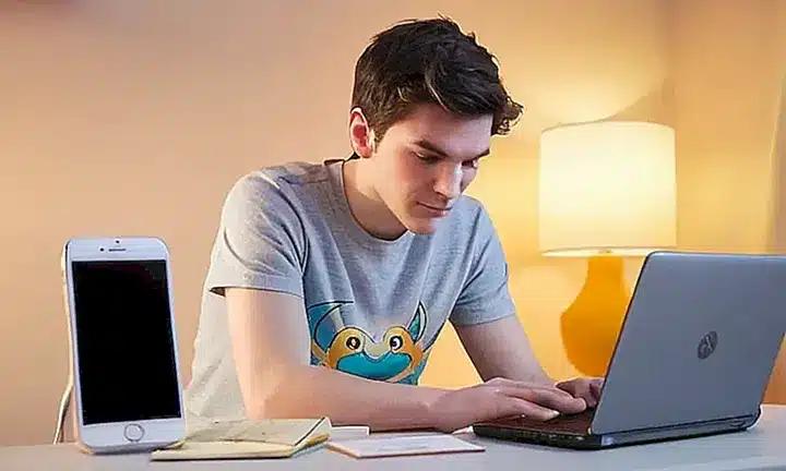Un joven dormitivo con un teléfono en la mano está sentado frente a su escritorio desordenado y jugando Pokémon Go en su computadora