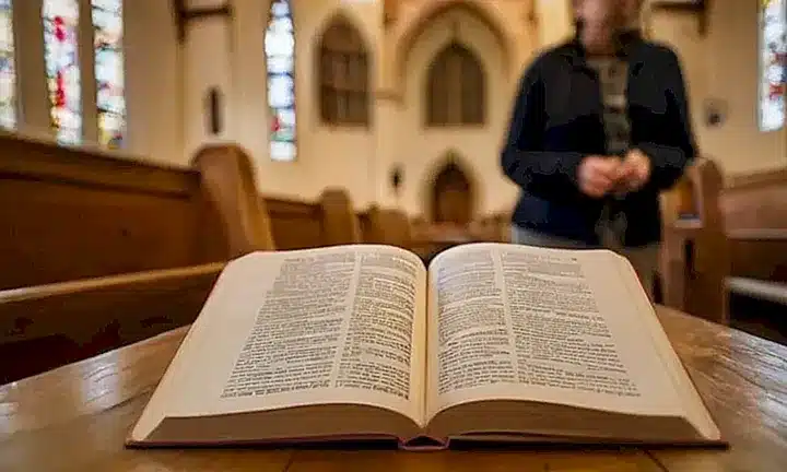 Una persona con gafas está sentada en un banco de madera en una iglesia oscura, leyendo con atención un libro