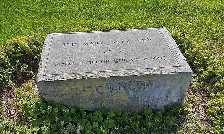 Una lápida con epitafio se encuentra en un entorno desgastado y deteriorado, bañado por la luz del sol