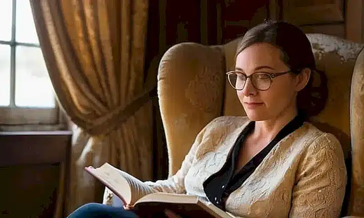 Una mujer con gafas sentada en un antiguo sillón lee un libro de cuero atado con notas manuscritas en una atmósfera tranquila y acogedora