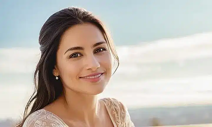 Una mujer sonriente con un vestido blanco y longitudes oscuras se encuentra en una serena escena rodeada de claridad suave