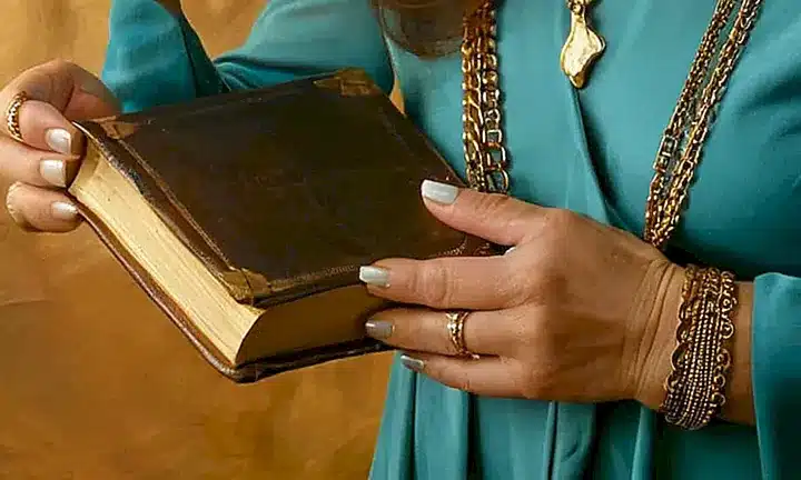 Una mujer madura sostiene un libro viejo con bordes dorados y sus dedos brillan con joyas de plata bajo el suave resplandor de la luz del estudio