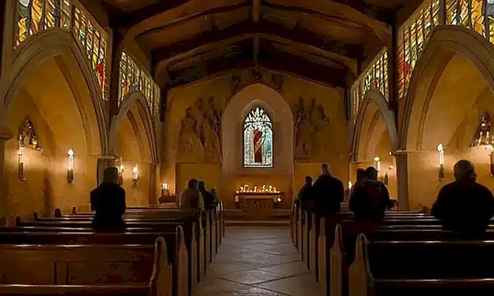 Un grupo de personas sientanse en silencio en una capilla oscura y elegante donde el sol cálido filtra a través de las ventanas de vidrio estrellado
