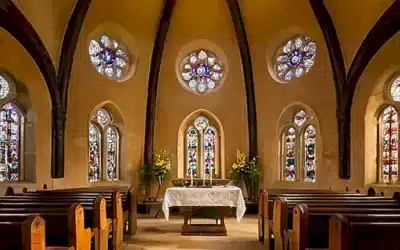 La iglesia tiene pilas y sillas antiguas rodeando un altar de piedra con luces claras y colores vibrantes creados por ventanas estampadas y olor a incienso