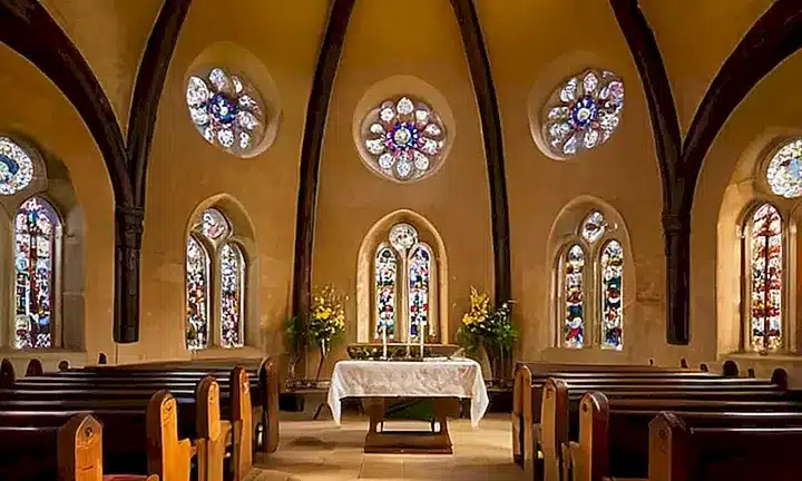 La iglesia tiene pilas y sillas antiguas rodeando un altar de piedra con luces claras y colores vibrantes creados por ventanas estampadas y olor a incienso