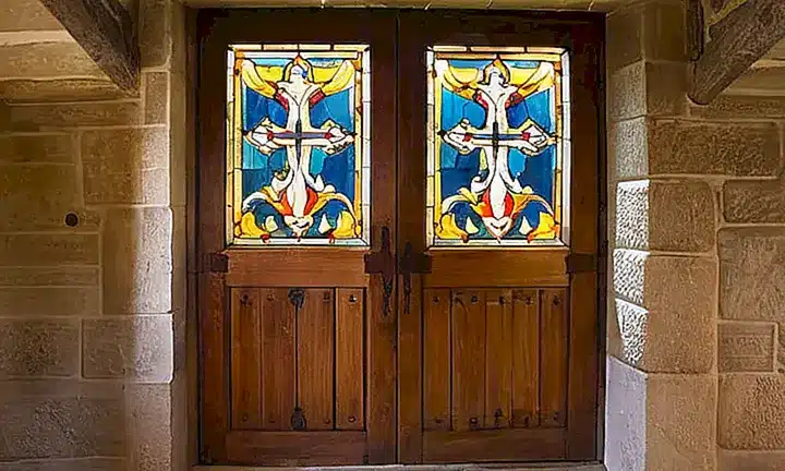 Una antigua iglesia tiene paredes de piedra gris, un puerta rústica con caracteres bíblicos y ventanas de vidrio coloreado que dejan pasar la luz natural