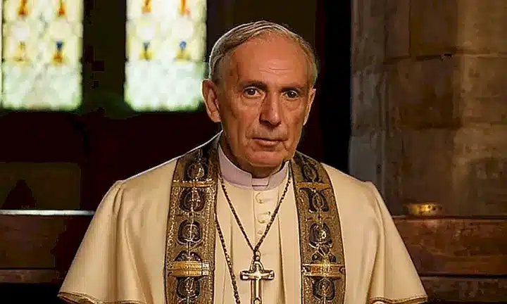 Un anciano obispo se encuentra bajo la luz tenue de velas, con atuendo formal y una cruz de oro que destaca su autoridad espiritual