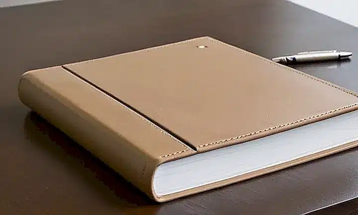 Un libro encuadernado en beige con letras plateadas embajadas se apoya sobre un escritorio gris claro, iluminado por una luz dorada que destaca la textura del mobiliario