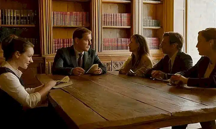 Un grupo de personas con expresiones faciales variadas se reúne alrededor de una mesa de madera vetada rodeado por objetos y fondos que agregan profundidad y interés visual a la escena
