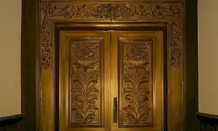 Una antigua puerta de madera ornamentada con detalles vegetales está parcialmente abierta, revelando un pasillo estrecho que conduce hacia una gran cúpula con frescos decadentes