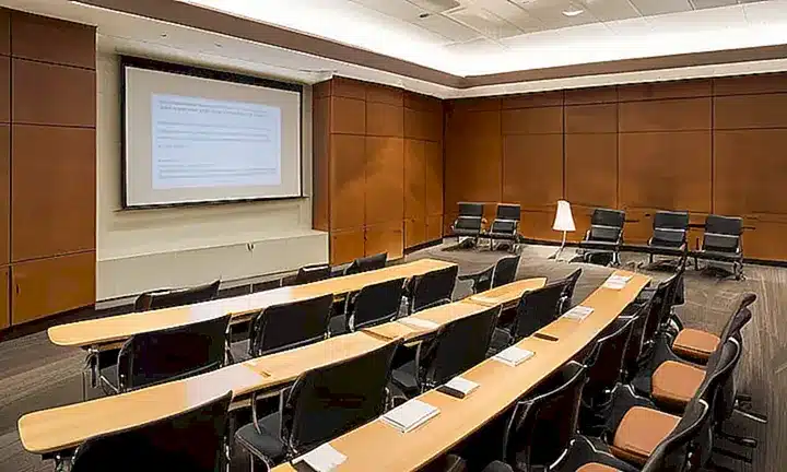 La habitación de conferencias cuenta con luces fluorescentes, escritorios y un panel de madera en el fondo decorados con imágenes de diagramas complejos