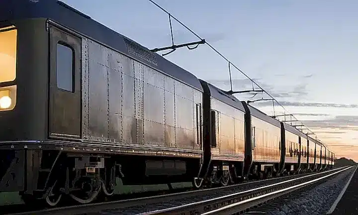 Una maqueta de un tren está colocada en una plataforma de madera con un fondo de computadora que muestra el entorno del juego Trainz Railroad Simulator 2019