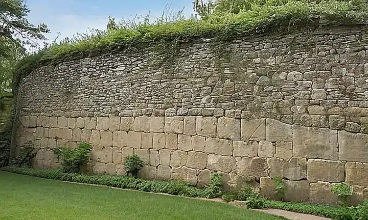 En la escena se muestra una mujer desalliñada con la mirada hacia el cielo, sentada detrás de un muro de piedra con luces que destacan su textura
