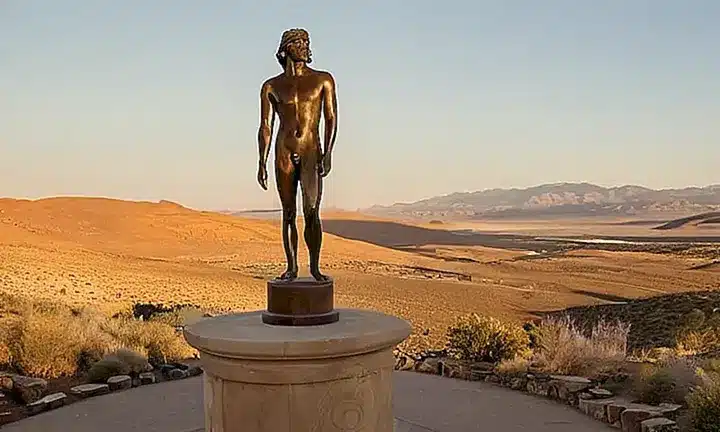 Un estatuaria en bronce con ropas desgastadas y accesorios de cuero descansa sobre un pedestal rodeado por un cielo claro desertico con sombras que se derraman desde una figura única