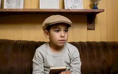 En una habitación simple y un poco desordenada se ve al niño sentado en un sofá frente a una librería y fotografías familiares con un café aún caliente