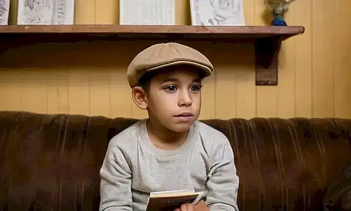 En una habitación simple y un poco desordenada se ve al niño sentado en un sofá frente a una librería y fotografías familiares con un café aún caliente