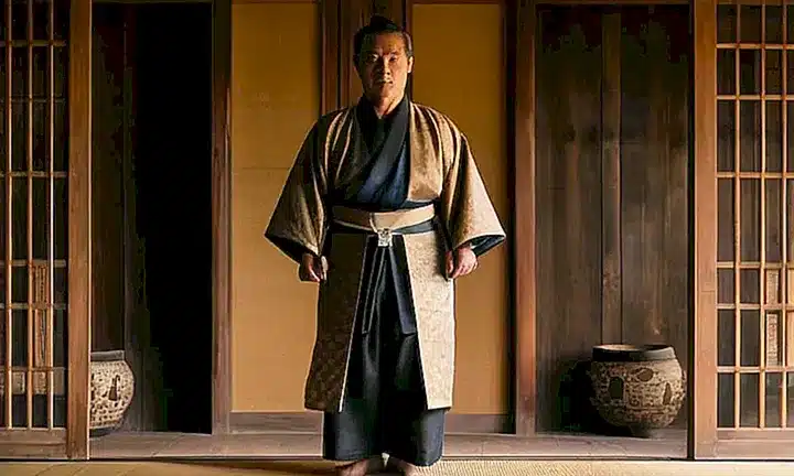 Un hombre de aspecto veterano se encuentra parado junto a una linterna de piedra, vestido con un atuendo japonés del siglo XVI