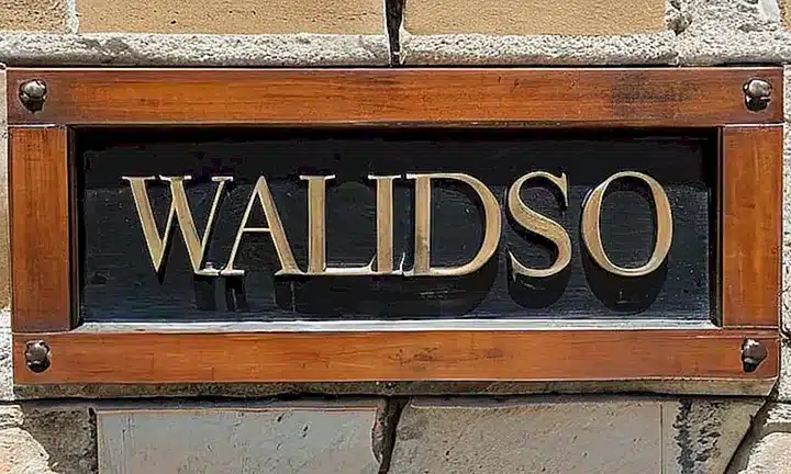 Un viejo rótulo de madera con letras grabadas cuelga en una pared de hormigón descolorida, frente a una lámpara que proyecta sombra y resalta las imperfecciones del lugar
