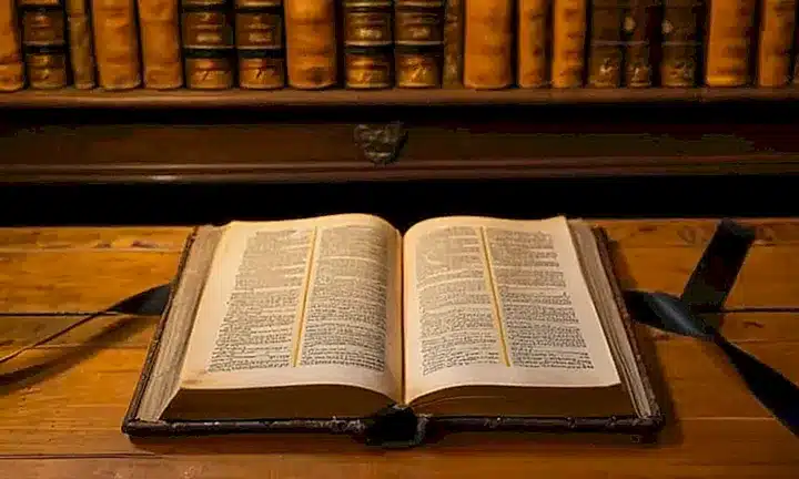 Un libro de Biblia antiguo y manuscritos se exhiben sobre un escritorio oscuro, iluminados por la luz suave de una vela