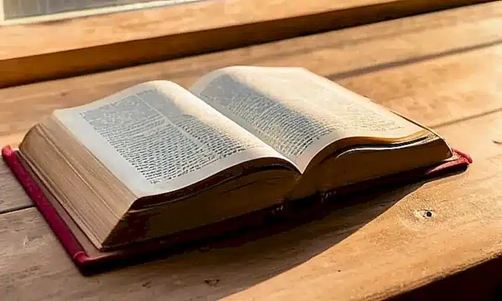 Una mano sostiene un antiguo Biblia en una mesa de madera sólida bajo la suave luz difusa que entra por la ventana