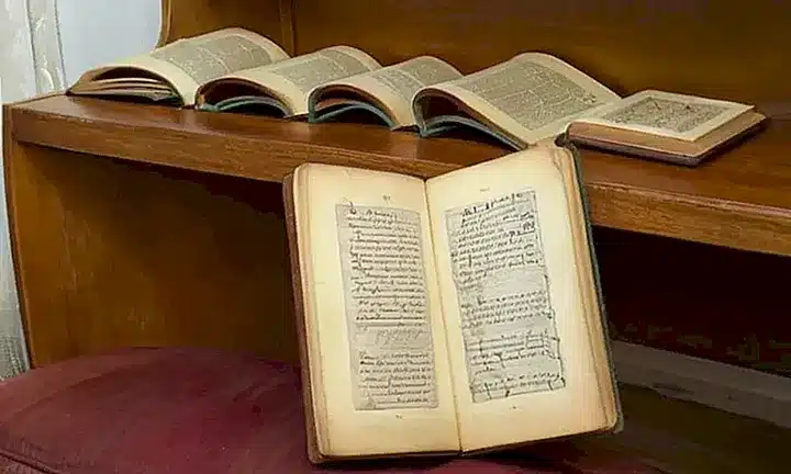 Una habitación silenciosa y acogedora donde una estantería con libros de cuero sostiene un bíblia antigua abierta sobre una mesa junto a una tumbona en la cual tres adultos charlan suavemente