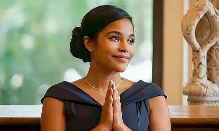 La joven mujer se sienta en oración con las manos juntas, rodeada de una luz dorada cálida, la ropa simple pero elegante y el fondo remoto de púlpitos que desvanecen en un distanciamiento suave