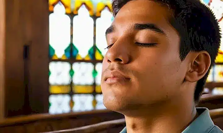 Un joven latino está en oración con expresión solemne, iluminado por el sol que pasa a través de las ventanas de vidrieras