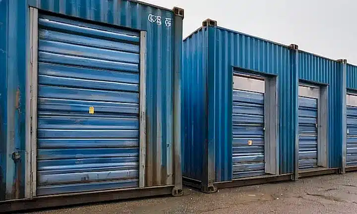 Un almacén de almacenamiento está compuesto por cajas metálicas oxidadas y deterioradas amontonadas entre sí, con entradas selladas por paneles de aluminio matizado desde las cuales se filtra la luz del sol