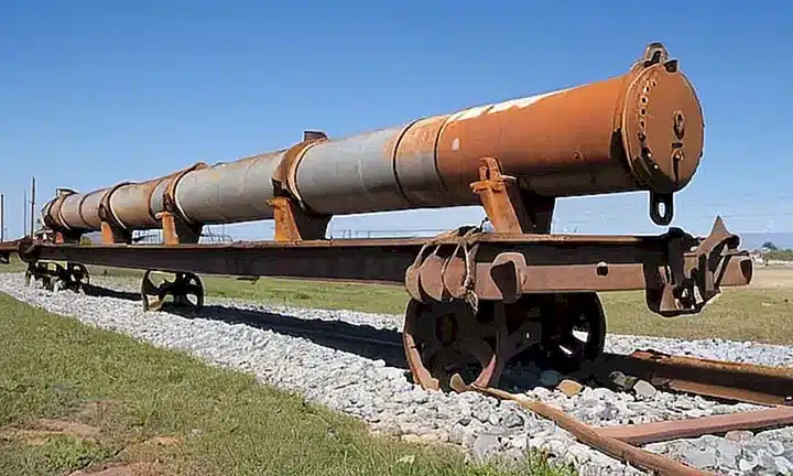 Un vagón de ferrocarril desgastado está parado en un andén cubierto de vías, rodeado de caños herrumbrados y cables eléctricos desteñidos
