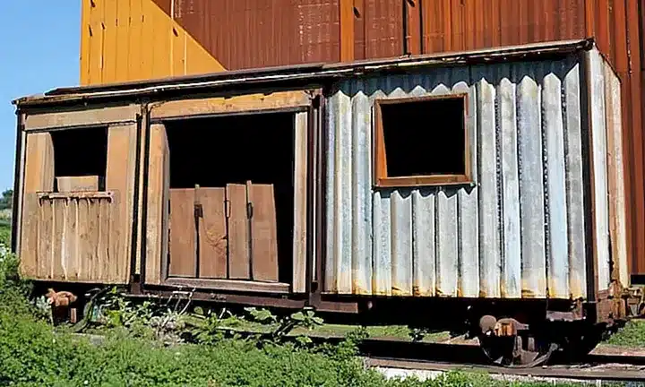 Un viejo depósito de ferrocarril está desmoronándose entre overgrowth y un edificio industrial en el fondo