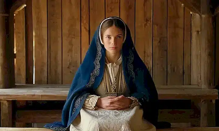 Una mujer del siglo XVII sentada en una bancal con vestimenta tradicional europea y un fondo iluminado por la luz solar que entra por una ventana de vidrio labrado