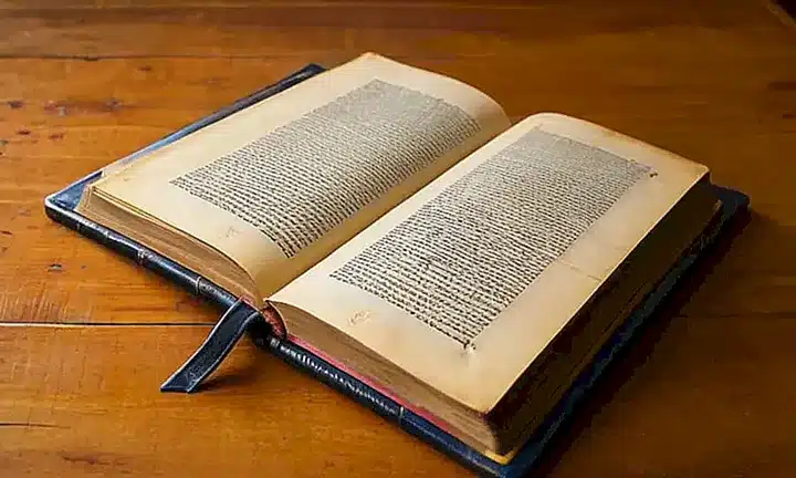 Un libro antiguo de cuero muestra signos de desgaste y uso, con páginas amarillentas, letras ornamentales en tonos oscuros y detalles físicos que dan cuenta de su edad